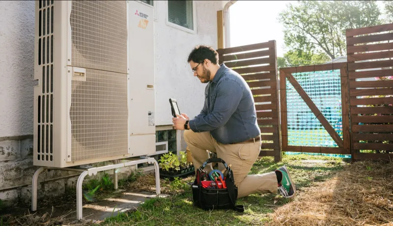 An image of a technician looking at an air source heat pump with his tool bag next to him.