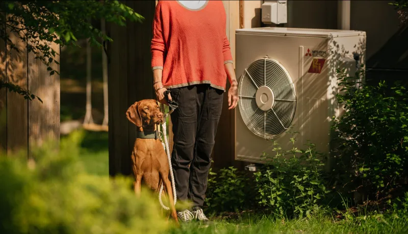 A person in a coral shirt stands between a dog and an air source heat pump.