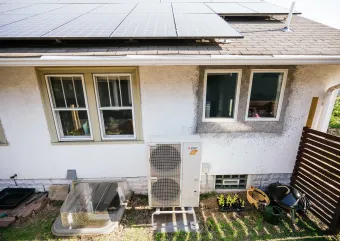 A white house with solar panels on the roof and a heat pump outside from afar.