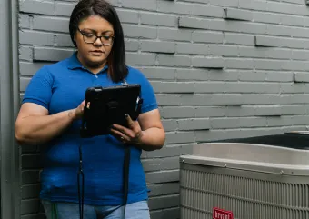A person stands holding an iPad during an inspection of a heat pump condenser.