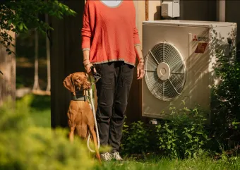 A person in a coral shirt stands between a dog and an air source heat pump.