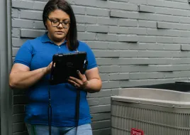 A person stands holding an iPad during an inspection of a heat pump condenser.