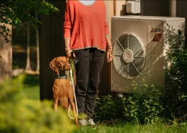 A person in a coral shirt stands between a dog and an air source heat pump.